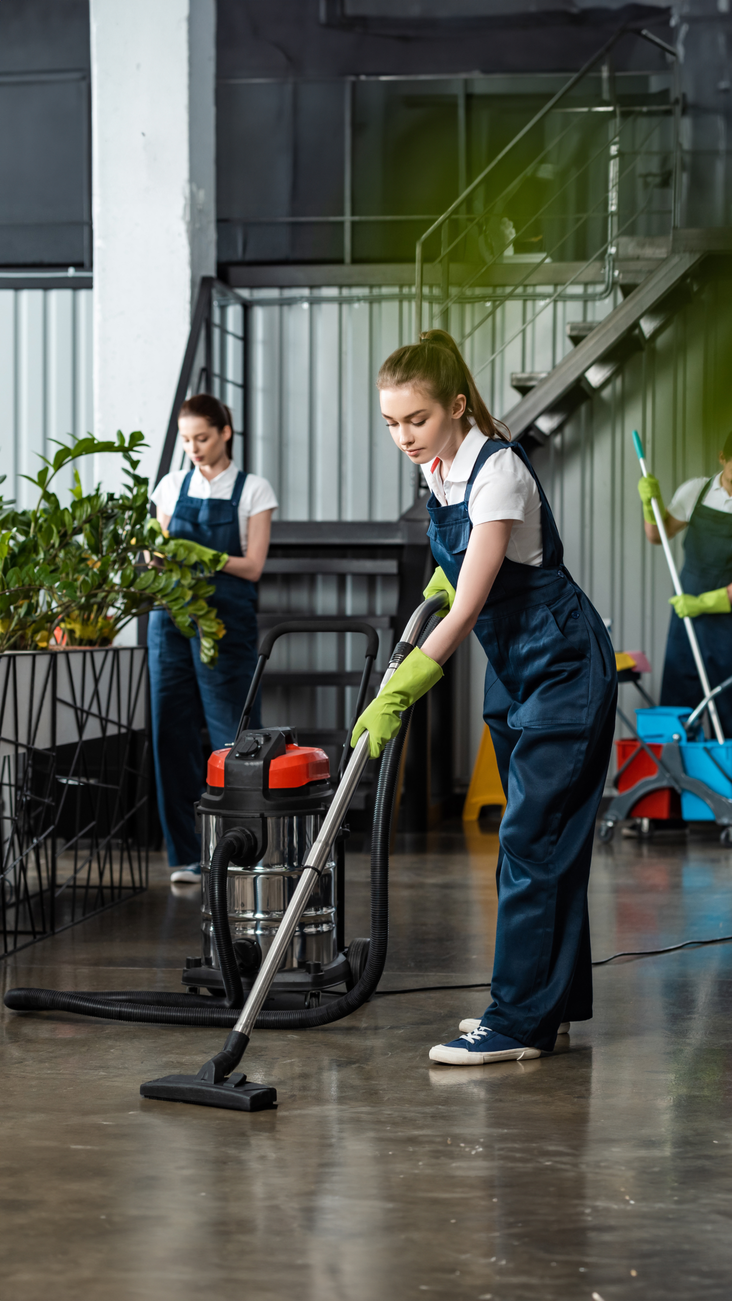 A young woman in a blue jumpsuit and green gloves is vacuuming a floor in a modern indoor space. Two other women are visible in the background, engaged in cleaning tasks. There are plants and cleaning supplies around.